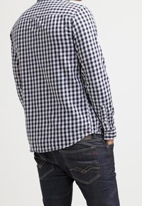 Camicia a maniche lunghe a scacchi blu e bianca con polsini a bottoni, indossata con jeans scuri caratterizzati da cuciture decorative e dettagli sulle tasche posteriori.