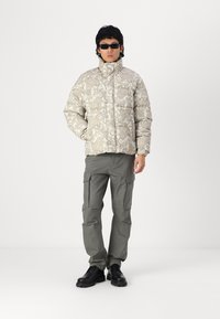 Padded jacket in light beige with paisley pattern, paired with gray cargo pants. Model also wears black sunglasses and black leather shoes.