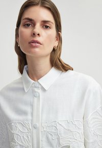 Camicia bianca con bottoni, realizzata in tessuto testurizzato e con un dettaglio di ricamo floreale sul davanti, caratterizzata da un colletto strutturato e bottoni chiari.