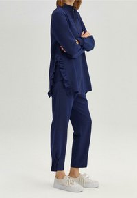 Donkerblauwe trainingspak met een ritsjasje met geplooide zijkant en bijpassende tapered broek. Het materiaal lijkt zacht en glad te zijn.