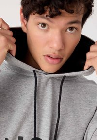 Sudadera gris con interior negro y cordones. La tela es suave y cuenta con un bolsillo frontal. Texto negro impreso visible en el frente.