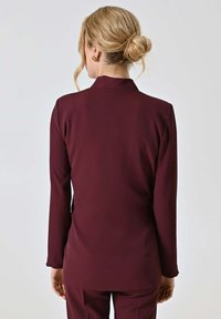 Blazer sartoriale color borgogna con maniche lunghe e collo alto, tessuto liscio, silhouette aderente, senza motivi o accenti visibili.