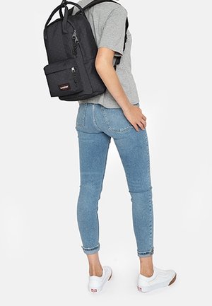 Henkilö, jolla on päällä vaaleansiniset farkut, valkoiset tennarit, harmaa t-paita ja musta reppu, jossa edessä vetoketjutasku, katsomatta kohti.