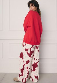 Hanorac roșu supradimensionat cu umeri căzuți, asortat cu pantaloni albi cu un model floral burgund. Texturi netede, croială lejeră.