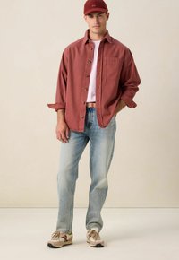 Rode overhemd met knopen, witte t-shirt eronder, lichte spijkerbroek, beige en bruine sneakers, rode pet, staand tegen een neutrale achtergrond.