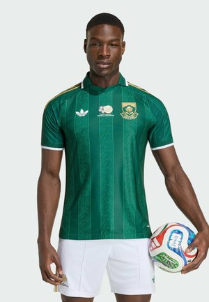 Atleta masculino con camiseta de fútbol sudafricana verde y pantalones cortos blancos, sosteniendo un balón de fútbol colorido con la mano derecha.