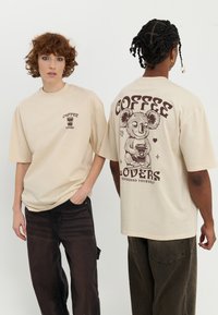 Beige oversized t-shirt lavet af bomuld, med front- og baggrafik med kaffe-tema, herunder et koala-design og tekst i brun.