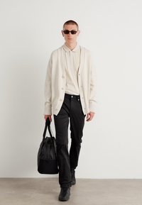Lichtbeige cardigan met knopen, witte polo, zwarte broek en donkere sneakers. Zwarte tote bag, zonnebril, minimalistisch ontwerp.