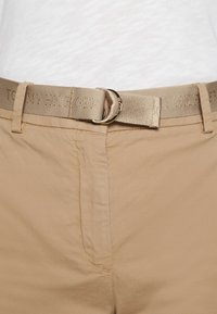 Beige stoffbelte med en glatt tekstur, med en rund metallspenne og preget "TOMMY HILFIGER" logo på båndet.