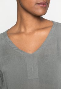 Blusa grigia a V con finitura testurizzata e dettagli di cucitura discreti sul collo. Il tessuto sembra morbido e leggero.