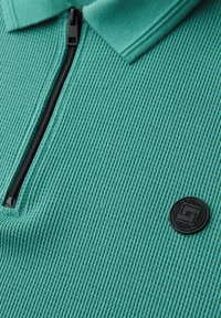 Gros plan sur une chemise en tissu texturé bleu sarcelle avec une fermeture éclair noire et un patch logo rond noir près du bas à droite.