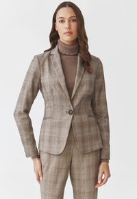 Hnědý kostkovaný blazer s přiléhavým střihem, jednořadým zapínáním na knoflík a klopami, kombinovaný s odpovídajícími kostkovanými kalhotami a hnědým rolákem.