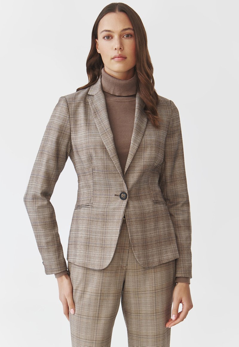 Hnědý kostkovaný blazer s přiléhavým střihem, jednořadým zapínáním na knoflík a klopami, kombinovaný s odpovídajícími kostkovanými kalhotami a hnědým rolákem.