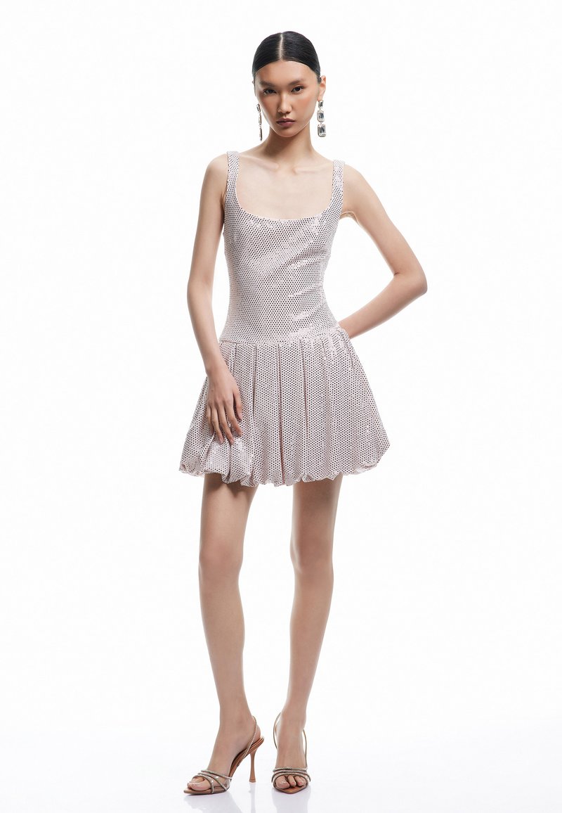 Robe mini ajustée et brillante en rose métallisé avec un décolleté carré et une jupe plissée. La modèle porte des boucles d'oreilles argentées et des sandales à talons.