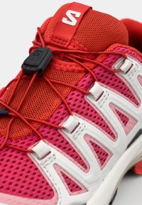 Scarpa sportiva rossa e rosa con sopra in rete, lacci elastici, dettagli bianchi, suola imbottita e suola in gomma con un motivo testurizzato.
