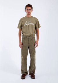 Oliwkowa koszulka z białym nadrukiem, zestawiona z oliwkowymi spodniami sztruksowymi i brązowymi butami. Dopasowany krój, krótkie rękawy, okrągły dekolt, teksturowana tkanina.