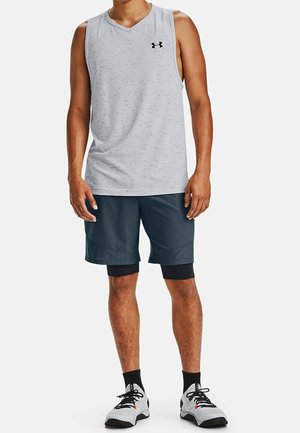 Mężczyzna w bezrękawniku w jasnoszarym kolorze, ciemnogranatowych szortach na czarnych spodenkach kompresyjnych, czarnych skarpetach i białych butach do biegania stoi.