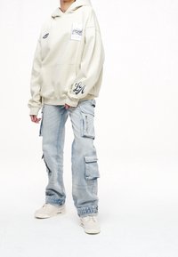 Beige hoodie med ficka och grafisk detalj. Ljust blå cargobyxor med blixtlås och flera fickor. Vita sneakers.