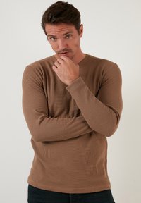 Braun gestreiftes Langarmshirt aus leichtem Stoff, mit Rundhalsausschnitt und enganliegenden Ärmeln. Lässig kombiniert mit dunklen Jeans.