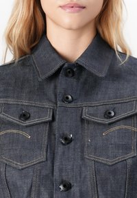 Close-up of a person wearing a dark denim jacket with black buttons and contrast stitching, showing collar and chest pockets.