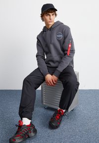 Sudadera gris con el logo de la NASA, bolsillo con cremallera y acento rojo, pantalones negros y zapatillas negras y rojas. Sentado sobre una caja metálica.