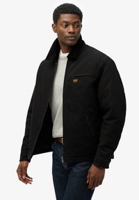 Veste bomber en coton noir avec fermeture éclair, col côtelé et poche poitrine. Portée par-dessus un pull gris, associée à un jean bleu.