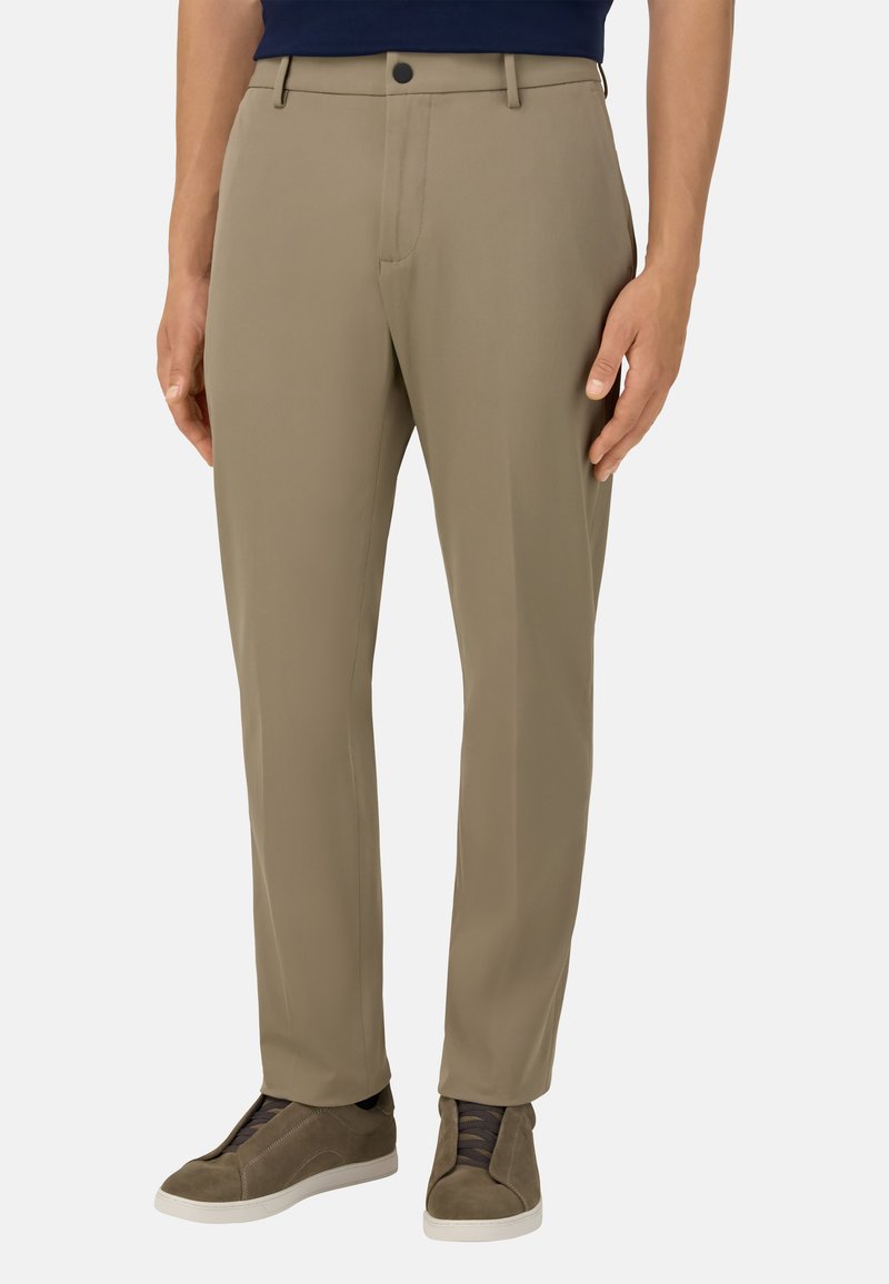 Béžové kalhoty na míru s hladkou texturou, s předním zipem, bočními kapsami a úzkým střihem, doplněné tmavými šněrovacími teniskami.