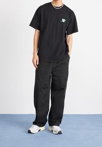 Camiseta de algodón negra con un gráfico verde en el pecho, combinada con pantalones holgados negros y zapatillas gris con detalles blancos.