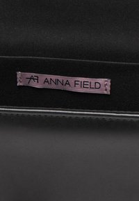 Fond en tissu noir avec une étiquette de vêtement violette cousue, affichant le nom de la marque "ANNA FIELD" en texte noir.