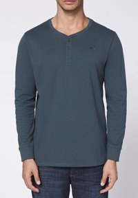 Langärmliges, dunkeltürkises Henley-Shirt aus Baumwolle mit einer Knopfleiste mit drei Knöpfen, abgerundetem Halsausschnitt und dezentem, gesticktem Logo auf der Brust.