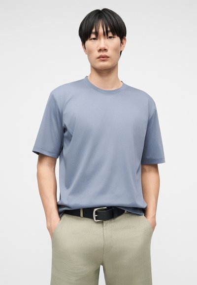 Νεαρός με μαύρα μαλλιά, φορώντας ένα ανοιχτό μπλε κοντομάνικο πουκάμισο μπα tucked μέσα σε μπεζ παντελόνι με μαύρη ζώνη, με τα χέρια στις τσέπες.