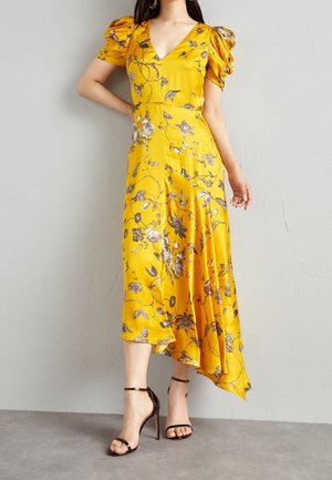 Mujer con vestido midi amarillo con flores, mangas abullonadas y sandalias negras de tacón alto, de pie contra una pared gris.