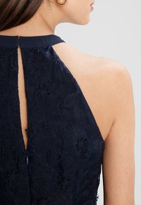 Navy blue dress featuring floral embroidery, high neckline, and a keyhole back with a zipper closure. Textured fabric adds depth.