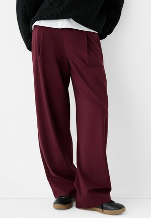 Person iført løstsiddende, mørk bourgognefarvede bukser, sort sweater, hvid skjorte og sorte sneakers med gummisåler foran en ensfarvet hvid baggrund.