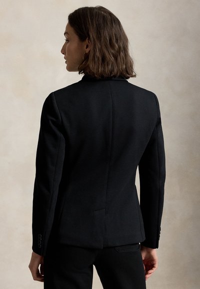 Donna con capelli castani fino alle spalle, indossa un blazer nero aderente e pantaloni neri, vista di schiena su uno sfondo chiaro e semplice.