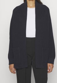 Marinblå stickad cardigan med långa ärmar, öppen front och framfickor. Har ett texturerat mönster samt ribbade mudd och nederkant.