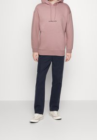 Lichtroze hoodie met een buidelzak en logo, gecombineerd met donkerblauwe broek en witte sneakers. Eenvoudig ontwerp met een relaxed fit.