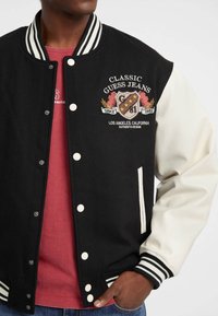 Mustvalged must ja valge varsity jakk riba servadega, nööpidega ja tikitud logoga. Seljas punane T-särk, mis sisaldab küljetaskuid.