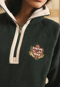 Tummanvihreä fleece-pusero, jossa on valkoinen vetoketjukaulus. Varustettu brodeeratulla kilpilogolla, jossa on kruunu, hevonen ja lehtikoristeet.