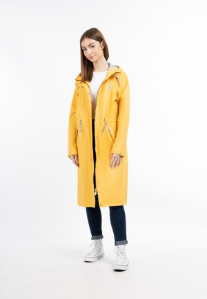 Impermeabile giallo con cappuccio, chiusura con zip, vita regolabile con cordino e due tasche laterali. Abbinato a jeans scuri e sneakers bianche.