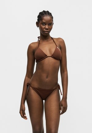 Femme portant un bikini triangle marron avec un col halter et un bas noué sur les côtés, debout devant un fond gris clair uni.
