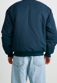 Giacca bomber blu navy realizzata in tessuto liscio e imbottito, con polsini e orlo a coste. Indossata con jeans in denim azzurro.