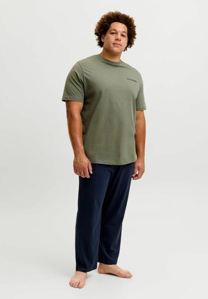 Homme aux cheveux bouclés portant une chemise verte à manches courtes et un pantalon bleu foncé, debout pieds nus sur un fond clair uni.
