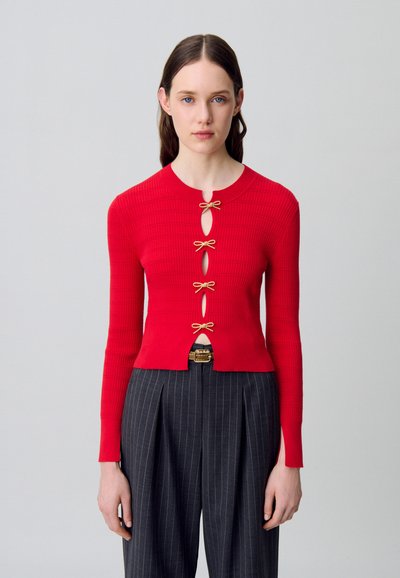 Cardigan rojo de canalé con mangas largas, que presenta recortes en forma de ojal y lazos en tono dorado como cierres. Llevado sobre pantalones de rayas de cintura alta.
