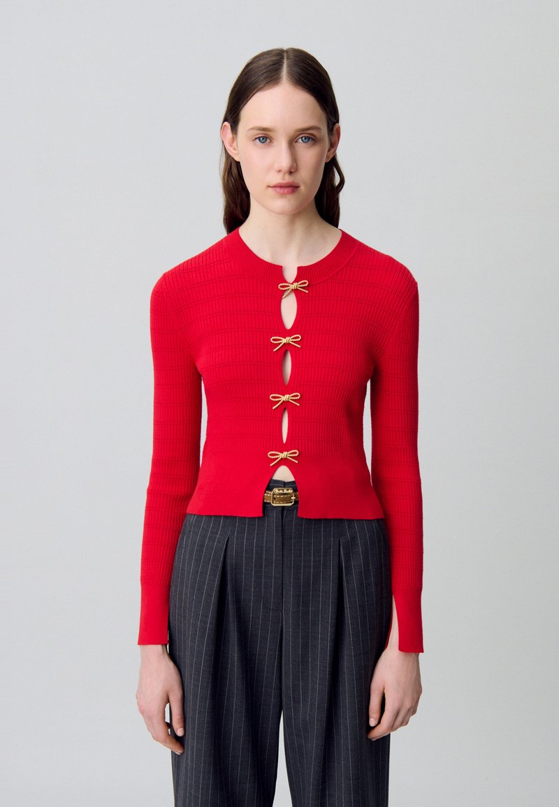 Cardigan rosso a costine con maniche lunghe, caratterizzato da aperture a forma di chiave e fiocchi in tonalità oro come chiusure. Indossato sopra pantaloni a righe verticali a vita alta.