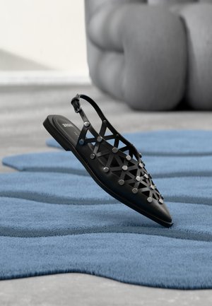 Zapato plano negro con punta puntiaguda y tira trasera, con cortes geométricos y tachuelas plateadas, sobre una superficie azul texturizada y fondo gris desenfocado.