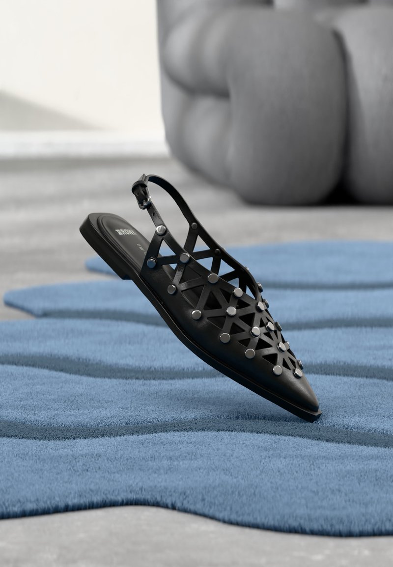 Μαύρο μυτερό επίπεδο παπούτσι slingback με γεωμετρικές εξαιρέσεις και ασημένια καρφιά σε μπλε υφή επιφάνεια και θολό γκρι φόντο.