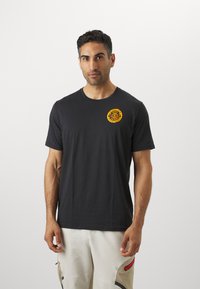 Schwarzes Baumwoll-T-Shirt mit rundem Halsausschnitt und kurzen Ärmeln. Ein auffälliges gelbes kreisförmiges Grafikmotiv ist auf der linken Brust aufgedruckt.