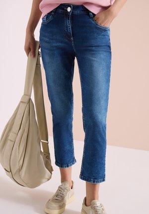 Persoon draagt cropped blauwe spijkerbroek, beige sneakers, houdt met één hand een beige schoudertas vast, andere hand in spijkerbroeken zak.