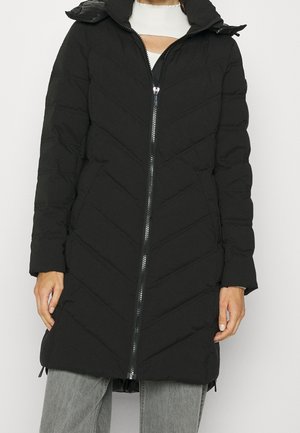 Czarny pikowany długi płaszcz z ukośnym przeszyciem, z przodu zamek błyskawiczny, wysoki kołnierz, noszony na białej bluzce i szarych dżinsach, modelka pokazana od szyi do ud.